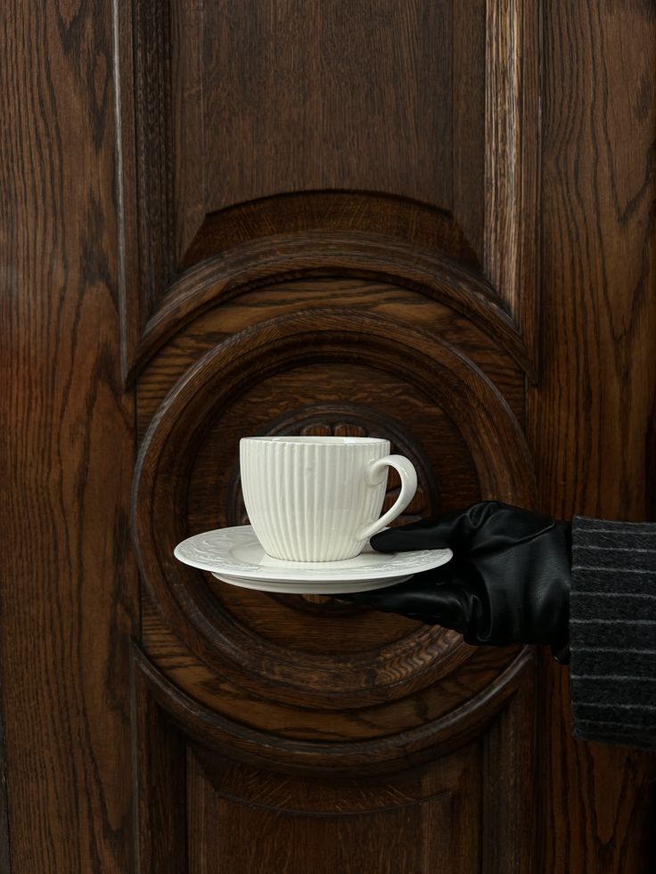 A gloved hand offering an espresso cup against a carved walnut door
